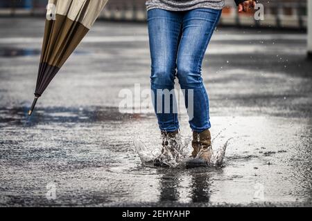 Saltare in pozzanghere e spruzzi d'acqua durante la pioggia. Donna che tiene l'ombrello e si trova in piedi sulla strada durante il tempo cattivo. Le gambe femminili indossano jeans e lea Foto Stock