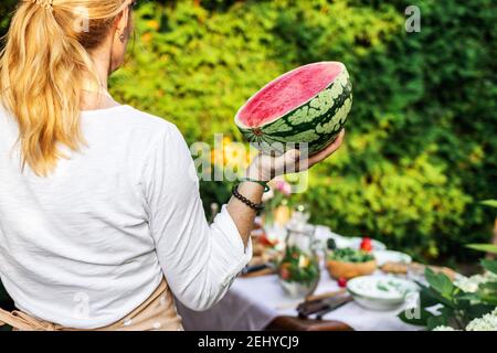 Donna che tiene il cocomero dimezzato. Preparazione del cibo per la festa estiva in giardino. Melone rosso maturo in mano femminile Foto Stock