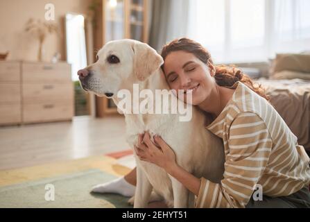 Ritratto dai toni caldi della giovane donna sorridente che abbraccia il cane mentre si siede sul pavimento e godendo del tempo con l'amante animale domestico, spazio di copia Foto Stock