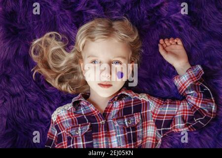 Giornata internazionale mondiale di consapevolezza delle malattie epilettiche. Carino bella bionda caucasica ragazza con piccolo viola porpora carta cuore sulla sua guancia sdraiata Foto Stock