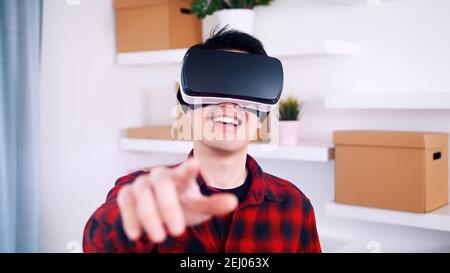 Felice giovane uomo sorridente mentre sperimenta la realtà virtuale e punta il dito verso la fotocamera. Foto di alta qualità Foto Stock