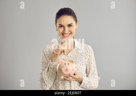 Donna guarda la fotocamera e mette una moneta in una banca piggy mentre si trova su uno sfondo grigio. Foto Stock