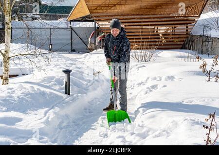 Il cortile è stato coperto di neve dopo una nevicata pesante. Un uomo pulisce i sentieri del giardino dalla neve in una giornata di sole in inverno. Il giardiniere si prende cura di th Foto Stock