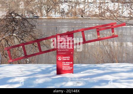 stazione a scala di soccorso in ghiaccio di colore rosso brillante con un avviso di pericolo Di ghiaccio sottile in Inwood Hill Park a New York dopo una tempesta di neve con terreno coperto di neve Foto Stock