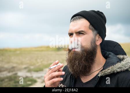 Uomo Barbuto il fumo di sigarette, bello hipster ritratto con faccia grave ha una lunga barba in hat outdoor su sfondo naturale Foto Stock