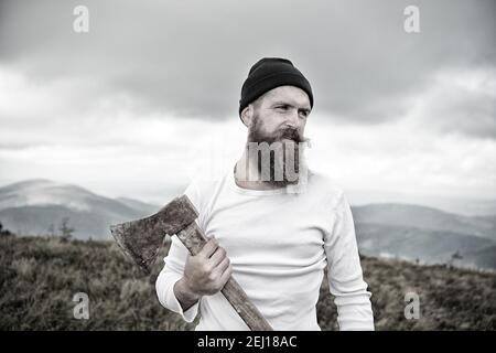 hipster. uomo bearded, barba lunga, brutale hipster caucasico con baffi tiene l'ascia con il viso serio sulla cima di montagna con cielo nuvoloso, ragazzo sraduto con i capelli alla moda ottenere barba taglio su sfondo naturale Foto Stock