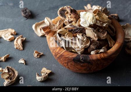 Primo piano immagine ad angolo isolata di una ciotola rustica di legno riempita con pezzi di funghi tagliati e secchi. Il recipiente si trova su una superficie di pietra scura. C'è un AS Foto Stock