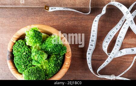 Immagine di una ciotola di legno di fiori freschi di broccoli e un nastro di misurazione fianco a fianco sulla superficie di legno. Immagine concettuale per dieta, fitness, noi Foto Stock