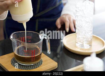 Il barista asiatico spreme il latte condensato da una bottiglia di plastica in una tazza per la misurazione del caffè. Atmosfera mattutina in una caffetteria. Foto Stock