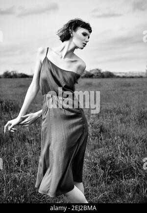 Foto di moda all'aperto: Bella ragazza triste nel campo. Ritratto di bella giovane donna in abito. Bianco e nero. Effetto grana pellicola Foto Stock