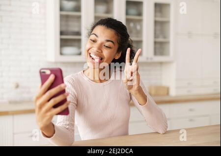 Teen biraciale divertente con i capelli scuri e la pelle di bronzo che mostra la lingua e scattando foto sul telefono, facendo un selfie, mostrando un segno del pezzo, a casa nel Foto Stock