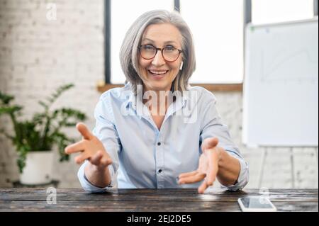 Felice amichevole senior insegnante manager con un sorriso piacevole in auricolari e occhiali guardando la macchina fotografica, seduta alla scrivania, gesturing e. Foto Stock