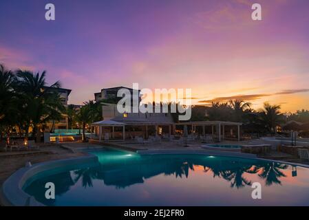 Splendido hotel con piscina all'alba, nei raggi dorati del sole. Riflessione nell'acqua blu delle nuvole rosa. Foto Stock