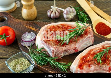 Pezzi di bistecca di maiale crudo con spezie ed erbe rosmarino, sale e pepe su tavola di legno su sfondo di legno in stile rustico Foto Stock