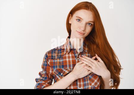 Giovane donna premurosa e sentita che tiene le mani a cuore, ringraziando per qualcosa, in piedi su sfondo bianco Foto Stock