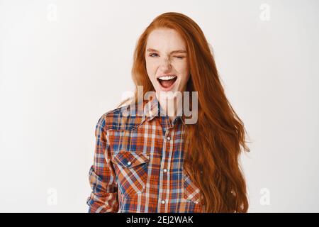 Ragazza attraente con i capelli di zenzero e gli occhi blu che si inocchiano alla macchina fotografica assuringly, in piedi su sfondo bianco Foto Stock