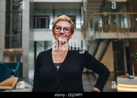 Ritratto di una donna d'affari senior di successo nell'edificio aziendale. Donna sicura con occhiali che guardano la macchina fotografica e sorridono. Foto Stock