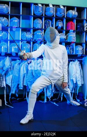 Giovane ragazzo in un vestito da scherma con una spada in mano, su uno sfondo blu, luce al neon. L'atleta si allena. Sport, gioventù, stile di vita sano. Foto Stock