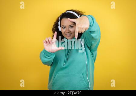 Giovane afroamericana che indossa abiti da palestra e utilizza le cuffie sorridente rendendo cornice con mani e dita con volto felice Foto Stock