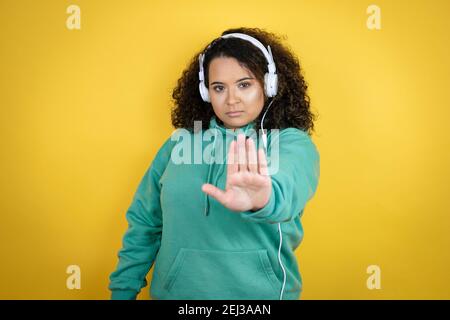 Giovane afroamericana ragazza indossando abiti da palestra e utilizzando le cuffie serio e smettere di cantare con il palmo della mano. Foto Stock