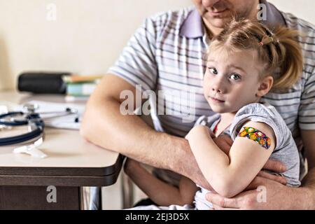 Vaccinazione di una bambina nelle braccia di suo padre nell'ufficio del medico dal coronavirus. Divertente cerotto adesivo per bambini. Vaccino contro covid-1 Foto Stock