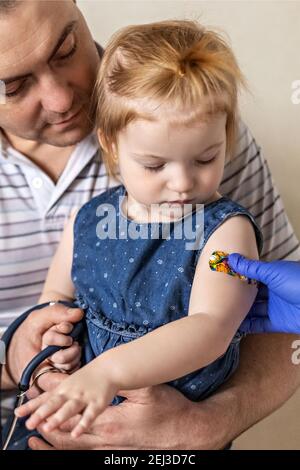 Vaccinazione di una bambina nelle braccia di suo padre nell'ufficio del medico dal coronavirus. Divertente cerotto adesivo per bambini. Vaccino contro covid-1 Foto Stock
