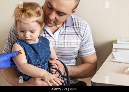 Vaccinazione di una bambina nelle braccia di suo padre nell'ufficio del medico dal coronavirus. Divertente cerotto adesivo per bambini. Vaccino contro covid-1 Foto Stock