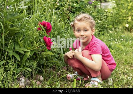 Una bambina si siede sorridente ad un letto di fiori nel giardino delle peonie rosa. Foto Stock