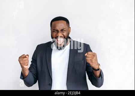 Emozionale eccitato afroamericano imprenditore maschile, freelancer o studente in abito elegante, in piedi su sfondo isolato, felice di vincere o notizie inaspettate Foto Stock