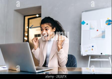 Entusiasta soddisfatta freelancer o studente afro-americano si siede in ufficio, guardando felicemente il portatile, ha ottenuto buone notizie, gioisce di buon affare Foto Stock