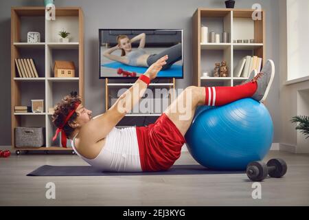 Divertente uomo grasso guardare la lezione di sport TV e fare fisico esercitarsi con la sfera di idoneità Foto Stock