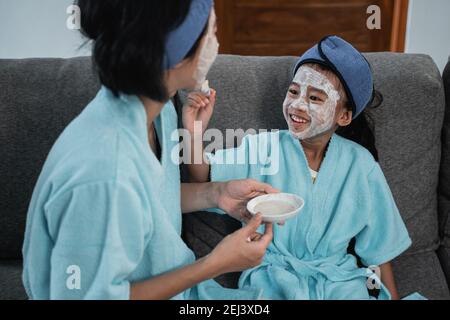 primo piano di una bambina che tiene una crema facciale spazzolare il viso della madre mentre si siede sul divano a casa Foto Stock