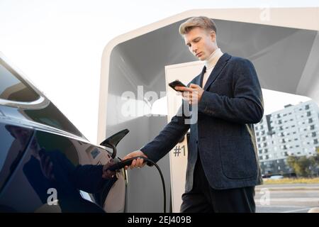 Giovane uomo d'affari caucasico piacevole usa lo smartphone durante l'attesa ricarica della batteria nella sua moderna auto elettrica su via Foto Stock