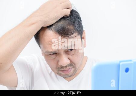 Un uomo di mezza età splende nello specchio e si preoccupa di perdita di capelli o grigio dei capelli isolato uno sfondo bianco, concetto di assistenza sanitaria Foto Stock