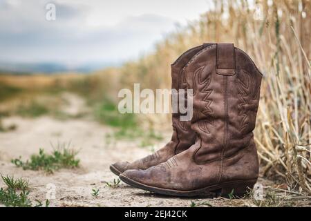 Stivale Cowboy su strada sterrata accanto al campo di grano. Vecchie scarpe in pelle usurate all'aperto. Stile retrò ancora vita di calzature in scena rurale Foto Stock