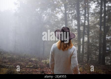 Moody atmosfera nella foresta. Donna che indossa maglia maglione e cappello è in piedi in nebbia a autunno freddo mattina. Godimento della natura Foto Stock