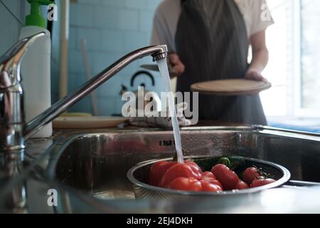 Verdure fresche sotto l'acqua corrente su un colino nel lavandino, sullo sfondo di una moglie con un coltello e un tagliere. Foto Stock