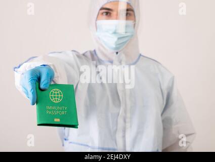 Medico in tuta protettiva di Hazmat che tiene il passaporto di immunità del coronavirus sullo sfondo dello studio luminoso, fuoco selettivo Foto Stock