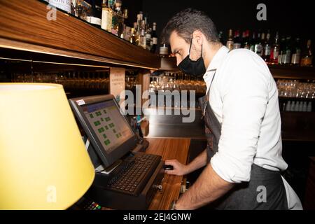 Barista focalizzato sul computer organizza gli ordini ai tavoli e. indossa una maschera di coronavirus Foto Stock