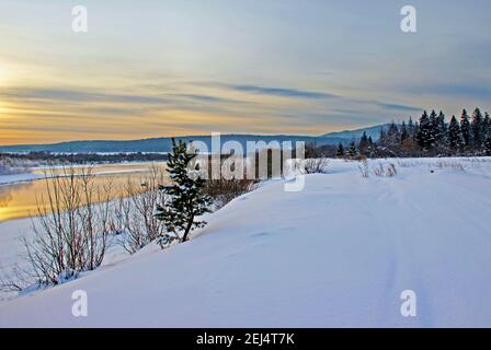 Vista su una riva del fiume innevata e montagne boscose al crepuscolo. Foto Stock