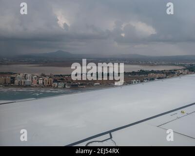 Splendida vista attraverso la finestra dell'aereo che si affaccia sulla città di Larnaca con edifici e lungomare, lago salato, mare blu con onde e montagne. Foto Stock