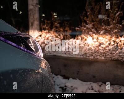 Faro auto bagnato con gocce d'acqua ghiacciate e neve che mette in evidenza i cespugli ricoperti di neve e marciapiede nel buio in una notte invernale. Foto Stock