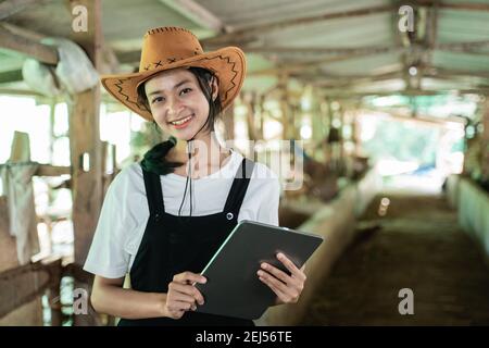 primo piano di una donna sorridente che indossa un cappello da cowboy portare un tablet digitale sullo sfondo di una mucca fattoria stabile Foto Stock