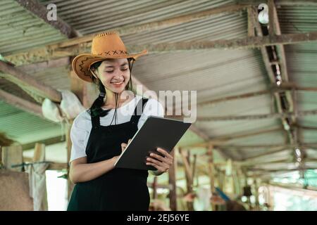 primo piano di una donna sorridente che indossa un cappello da cowboy mentre toccare lo schermo di un tablet digitale sullo sfondo di una stalla di mucca Foto Stock
