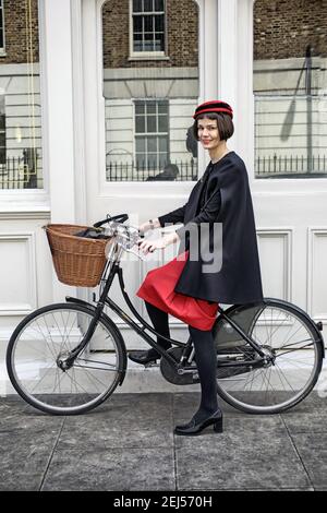 Giovane donna in abito rosso guida una bicicletta con cesto a Londra, Regno Unito Foto Stock