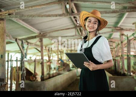 primo piano di una donna sorridente che indossa un cappello da cowboy guardando sulla fotocamera mentre si tocca uno schermo di un digitale tavoletta sullo sfondo di un allevamento di mucche stabile Foto Stock