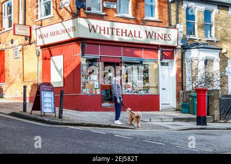 Un uomo con un cane su un cavo in attesa di attraversare la strada fuori Whitehall Wines, un locale fuori licenza a Londra del Nord, Regno Unito Foto Stock