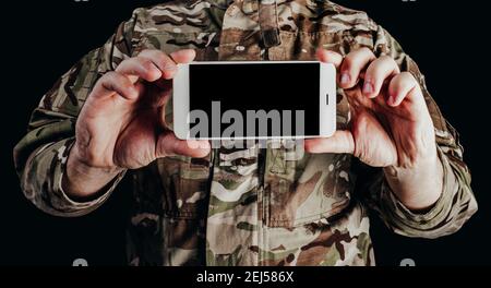 Foto del soldato in uniforme camuffata che tiene lo smartphone su sfondo nero. Foto Stock