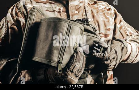 Foto del soldato in uniforme camuffata e guanti tattici che tengono la borsa delle gambe su sfondo nero. Foto Stock