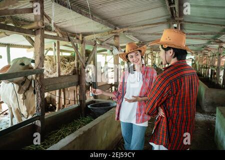 femmina coltivatore di mucca che indossa un cappello si alza chiacchierando con un partner maschile guardando le mucche nella fattoria mucca capanna Foto Stock